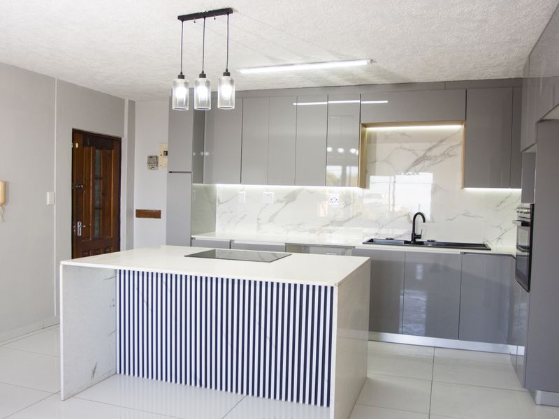 Modern kitchen featuring a central island with hob, sleek grey cabinetry, marble-look splashback, integrated oven, and contemporary pendant lighting.