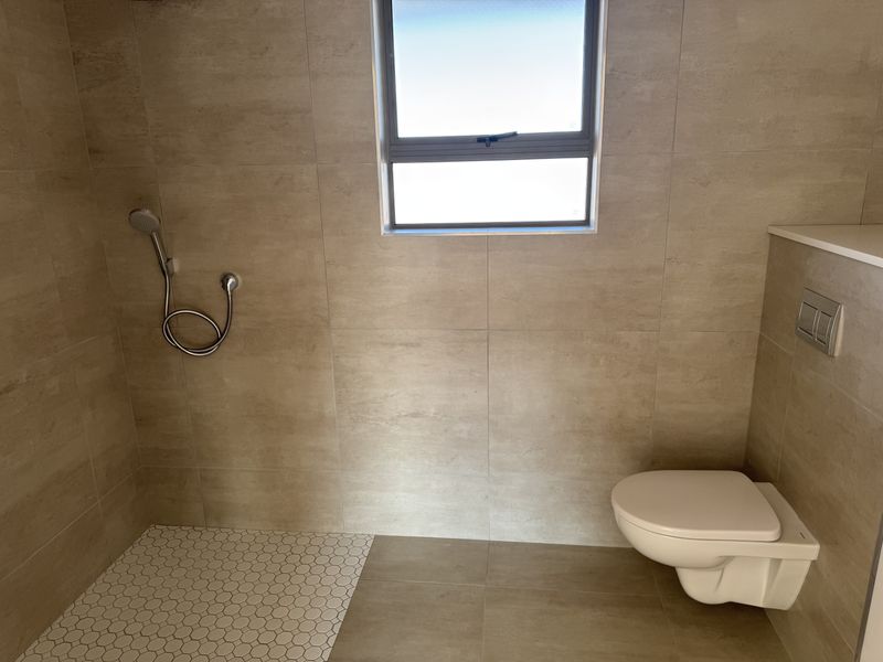 A modern, minimalist bathroom featuring a wall-mounted toilet, neutral tiled walls, and a walk-in shower area with a handheld shower fixture.