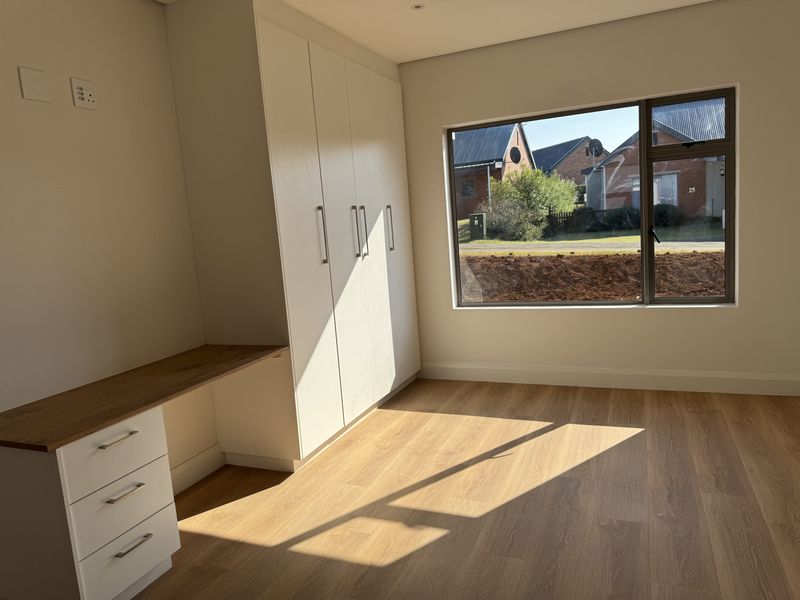 A bright, modern bedroom featuring built-in cupboards, a functional desk area, and high-quality laminate flooring with large windows providing natural light.