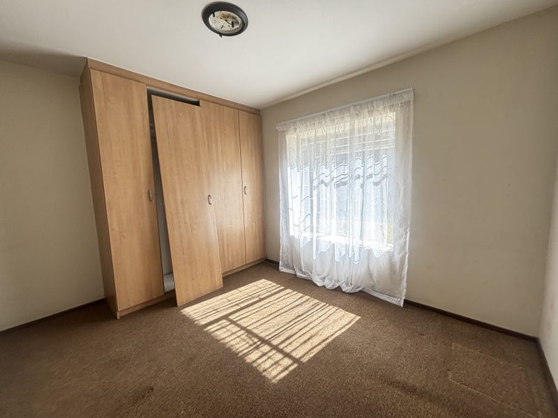 A vacant bedroom featuring built-in wooden cupboards, carpeted flooring, and a window with security bars, offering a blank canvas for renovation.
