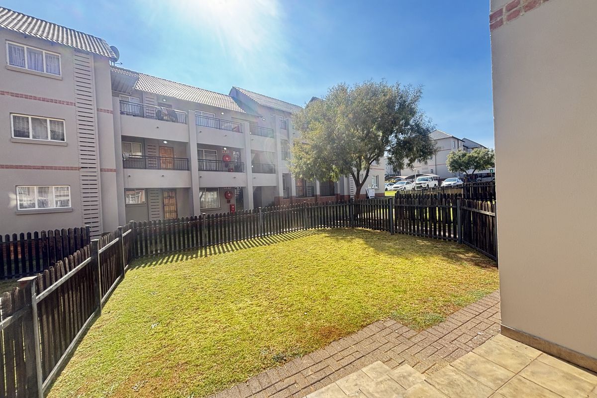 A private rear garden area featuring a paved patio, well-maintained lawn, and timber fencing, overlooking the apartment complex building.
