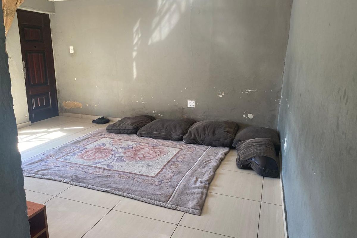 A sparsely furnished living room featuring tiled flooring and a solid wood entrance door, requiring cosmetic wall repairs and fresh paint.