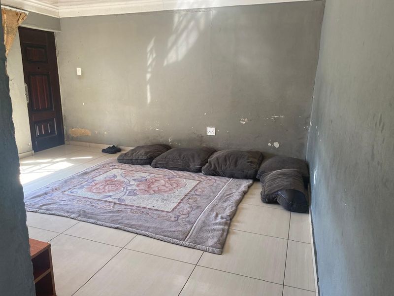 A sparsely furnished living room featuring tiled flooring and a solid wood entrance door, requiring cosmetic wall repairs and fresh paint.