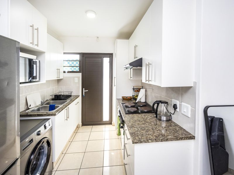 A functional galley-style kitchen featuring white cabinetry, granite-look laminate countertops, a built-in electric hob, and tiled flooring with direct access to the exterior.