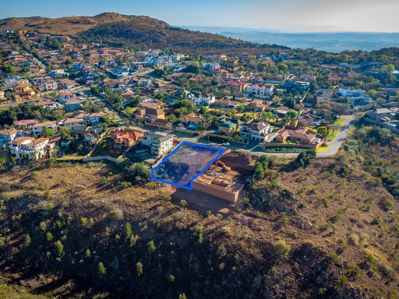 Aerial view of a vacant residential plot featuring a retaining wall and excavation work, situated in a developed hillside neighborhood.
