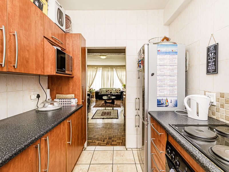 A functional galley-style kitchen featuring wood-look cabinetry, dark countertops, and tiled finishes, with a direct view into the adjacent living area.