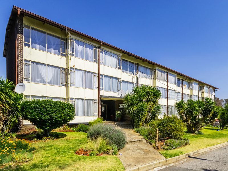 A multi-story apartment building exterior featuring large windows, a paved entrance walkway, and established garden landscaping.
