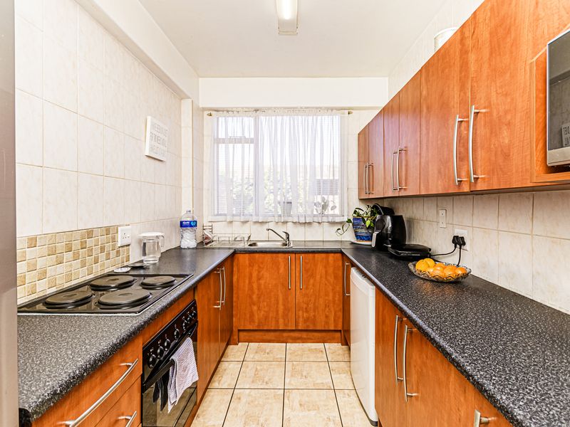 A functional galley-style kitchen featuring wood-finish cabinetry, dark laminate countertops, tiled backsplash, and integrated appliances including an electric hob and oven.