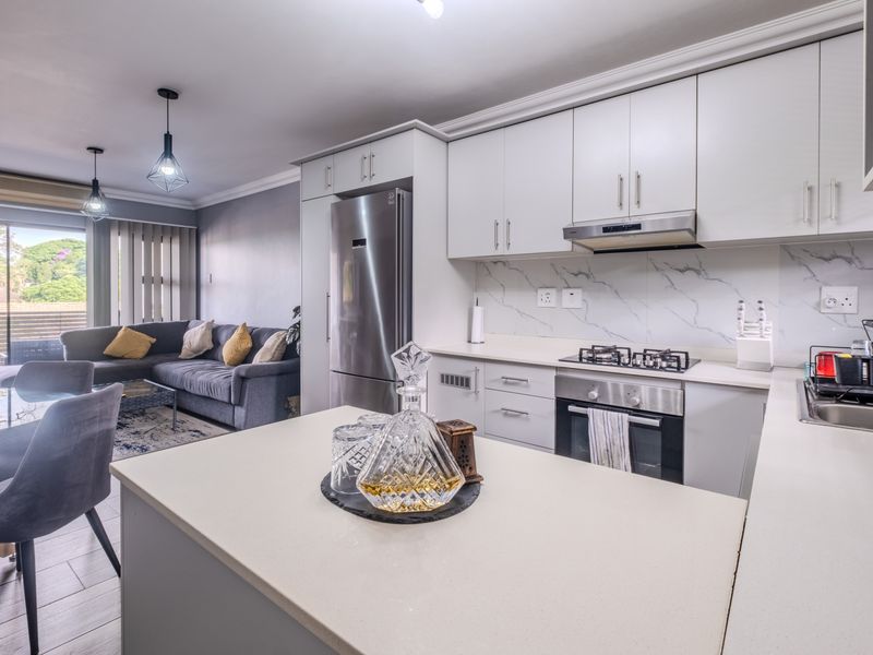 Modern open-plan kitchen and living area featuring sleek white cabinetry, stone countertops, integrated appliances, and direct access to a balcony.