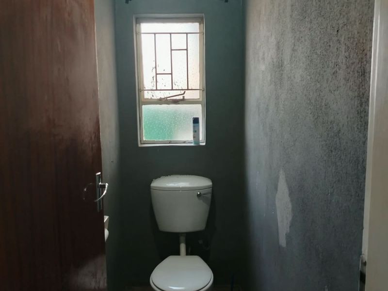 A small, functional bathroom featuring a standard toilet, tiled flooring, and a window with security bars, currently requiring cosmetic wall repairs and painting.