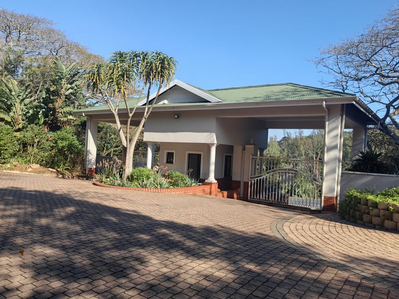 A well-maintained residential entrance featuring a paved driveway, covered portico, and secure metal gate set within a landscaped garden.