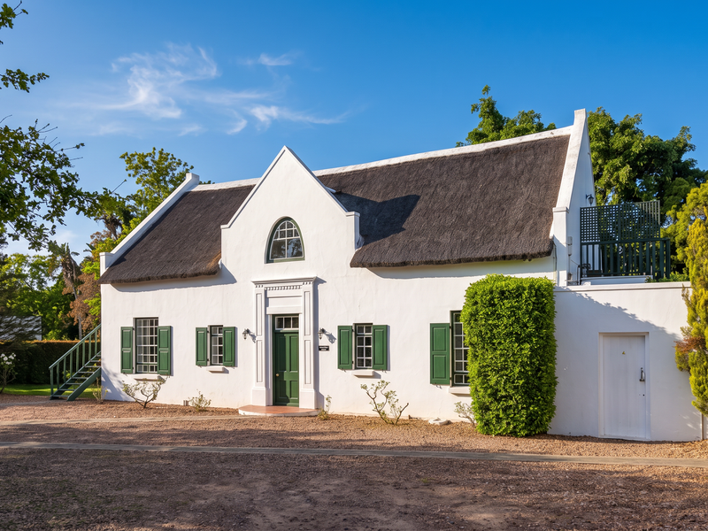 A charming Cape Dutch style guest house featuring a traditional thatched roof, white exterior walls, green timber shutters, and a well-maintained gravel driveway.