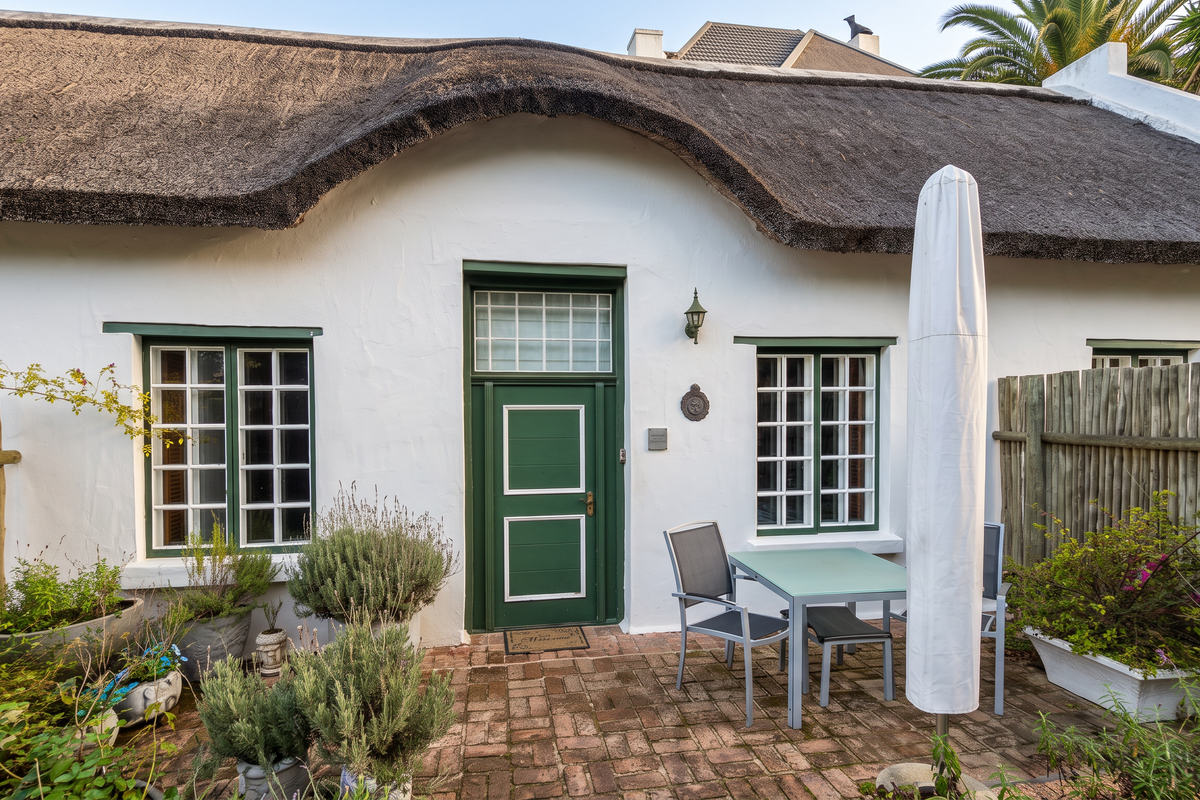 A charming guest house exterior featuring a traditional thatched roof, white-washed walls, and a private brick-paved patio area.