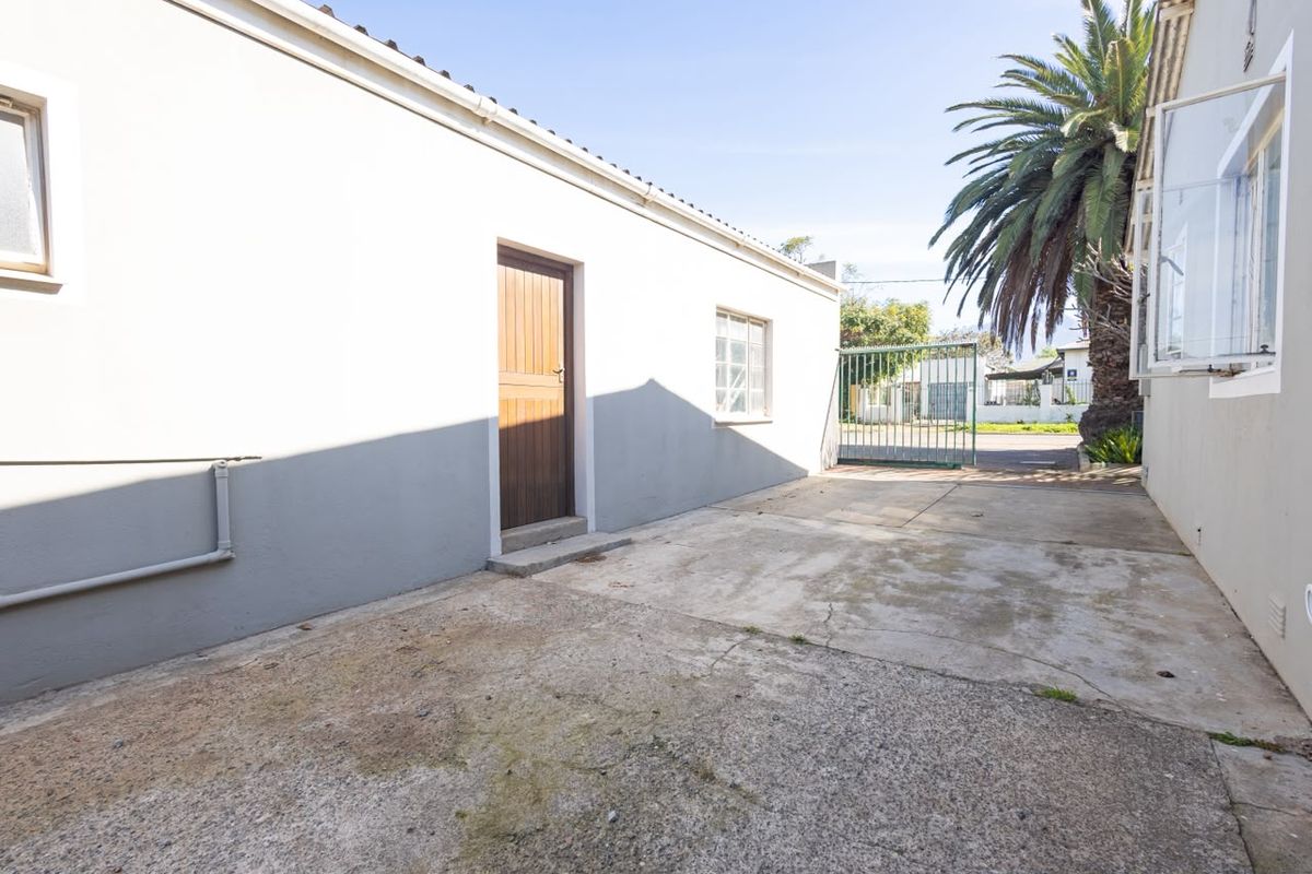 A rear exterior view showing a concrete driveway, a timber door entrance, and a metal security gate leading to the street.