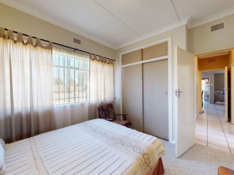 A bedroom featuring built-in sliding door wardrobes, security bars on the window, and carpeted flooring, with a view into the adjacent tiled hallway.