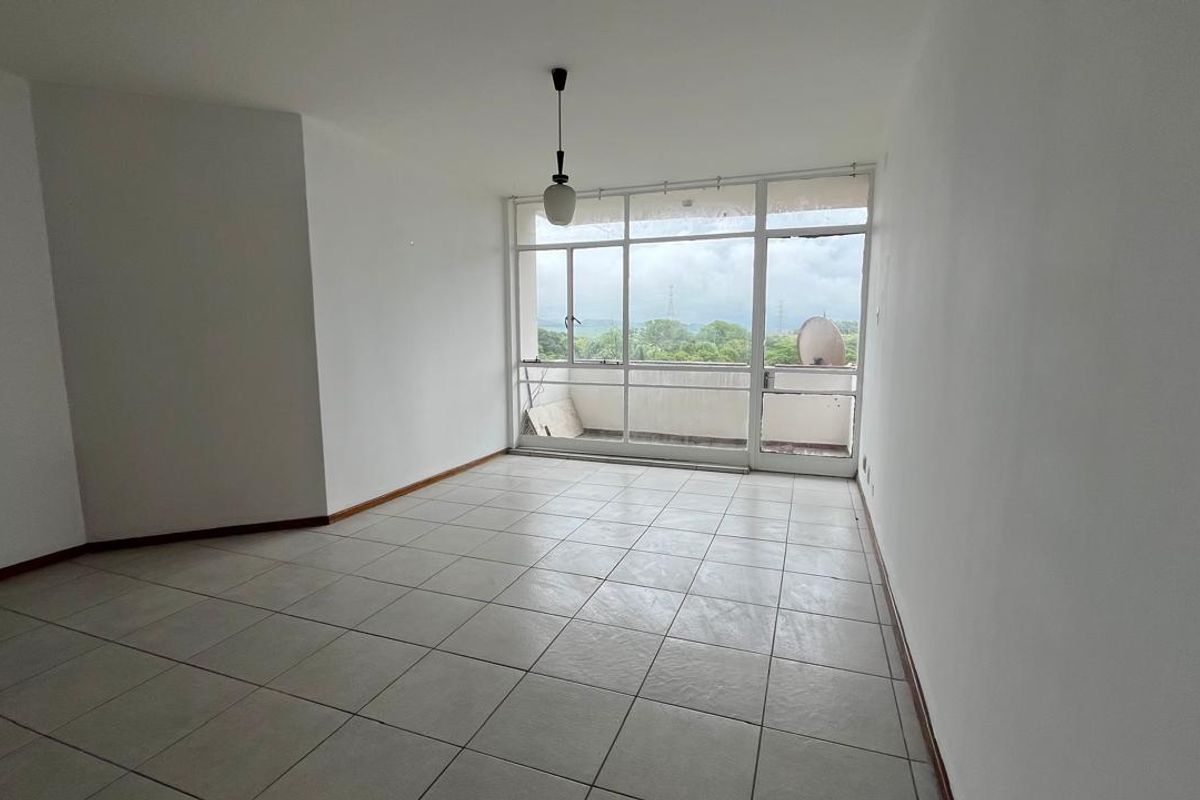 An empty, sunlit living area featuring neutral tiled flooring and large glass doors leading to an outdoor balcony with scenic views.