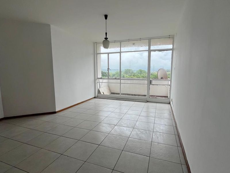 An empty, sunlit living area featuring neutral tiled flooring and large glass doors leading to an outdoor balcony with scenic views.