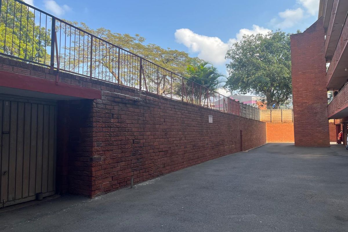 Exterior view of an apartment complex rear area featuring a brick retaining wall, metal safety railing, and paved driveway access to garages.