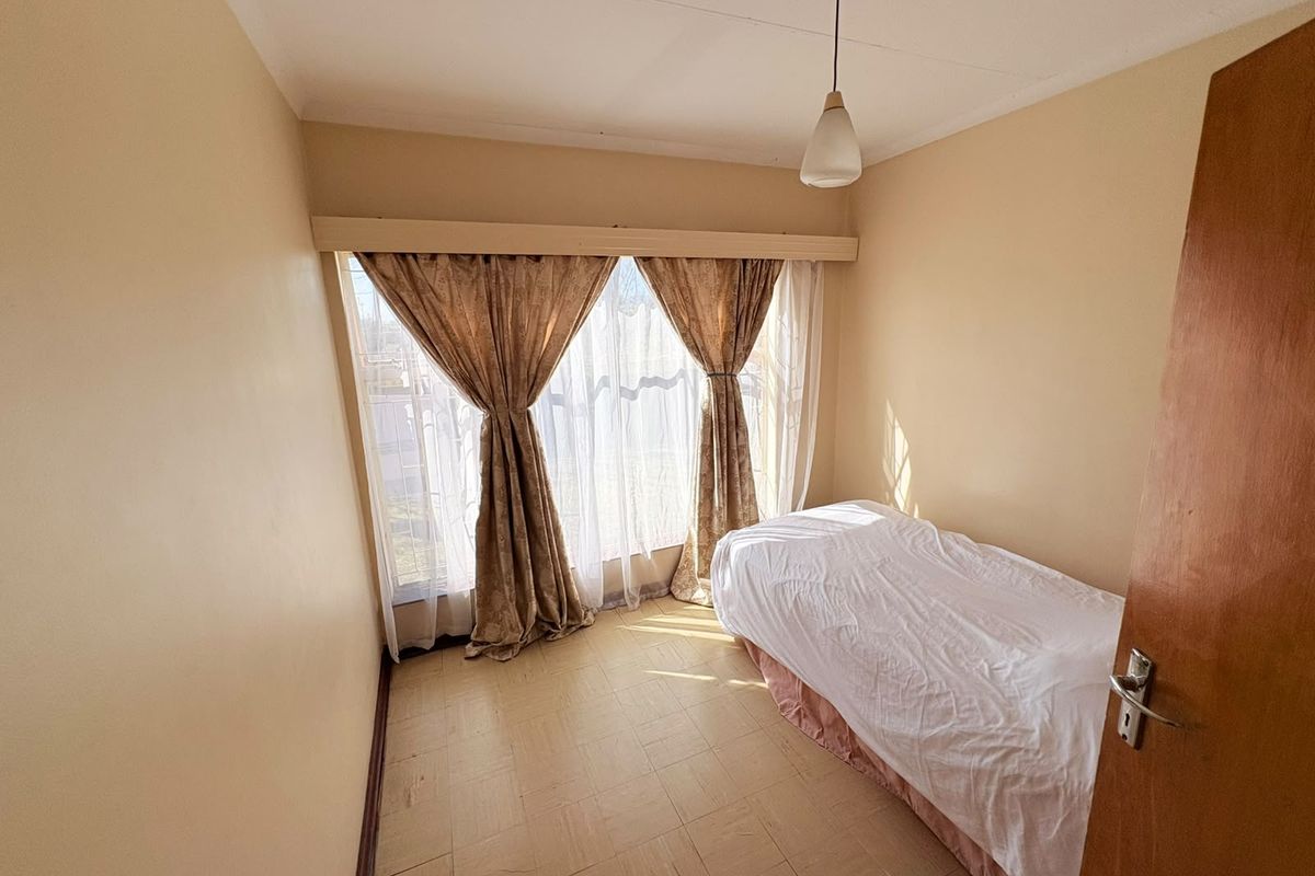 A compact bedroom featuring parquet flooring, a central pendant light, and a large window with a fitted curtain rail.