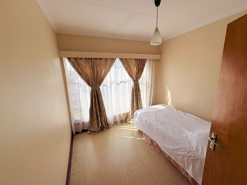 A compact bedroom featuring parquet flooring, a central pendant light, and a large window with a fitted curtain rail.