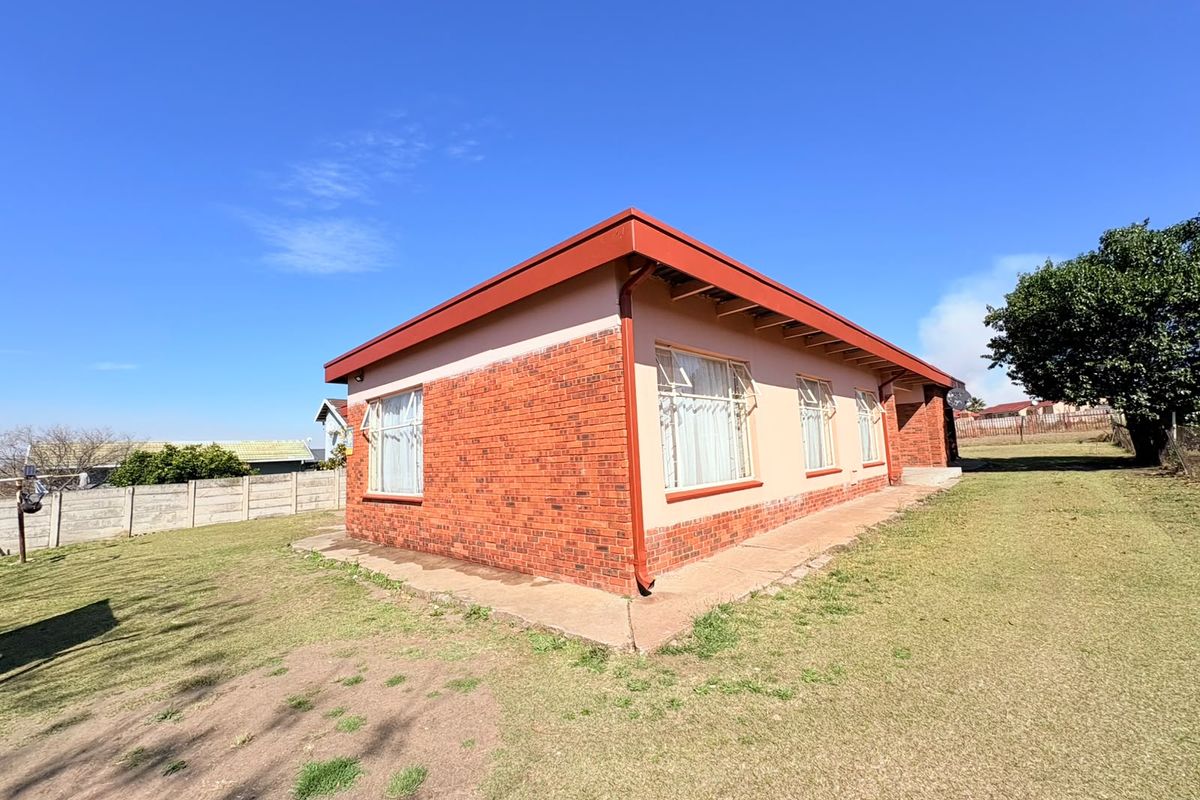 Rear view of a single-story brick house featuring a metal roof and concrete perimeter apron set on a large grassed plot.