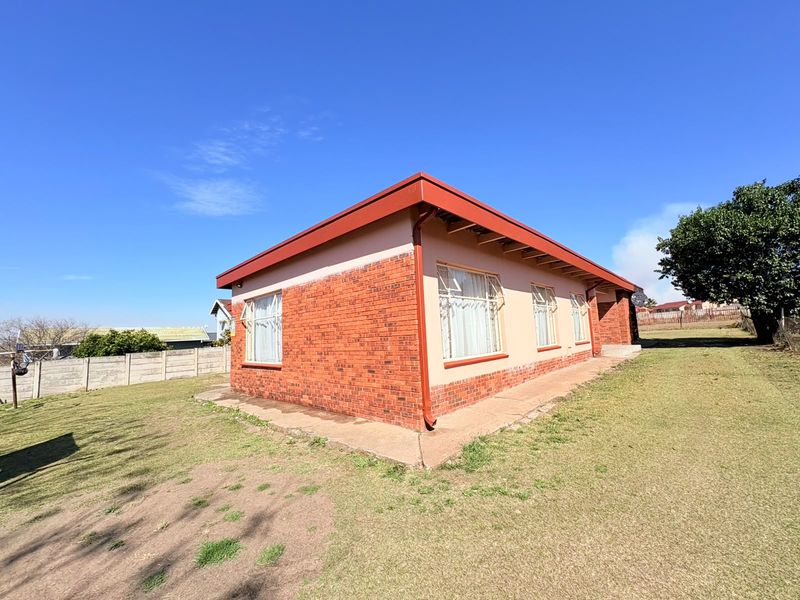 Rear view of a single-story brick house featuring a metal roof and concrete perimeter apron set on a large grassed plot.