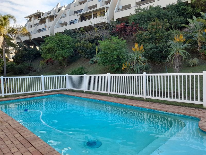 A well-maintained swimming pool area featuring brick coping and a white picket fence, set against a landscaped hillside with apartment building views.