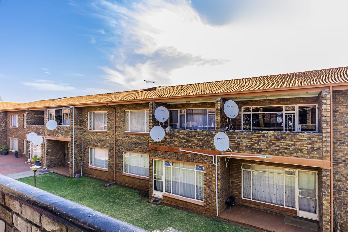 Rear view of a multi-story apartment complex featuring face brick construction, terracotta roof tiles, and ground-level patio access with established lawn.