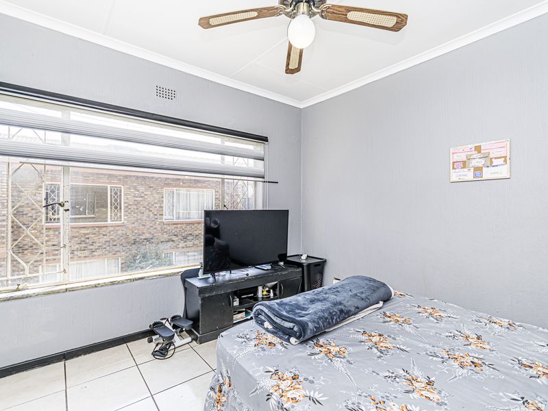 A bedroom featuring tiled flooring, a ceiling fan, and large windows with security bars and roller blinds.
