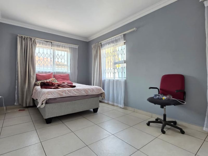 A bedroom featuring tiled flooring, neutral grey walls, and windows fitted with security bars, offering a functional and well-lit living space.