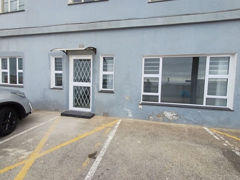 Front exterior view of the flat featuring a security gate, aluminium windows, and a paved parking area, showing signs of exterior wall damage.