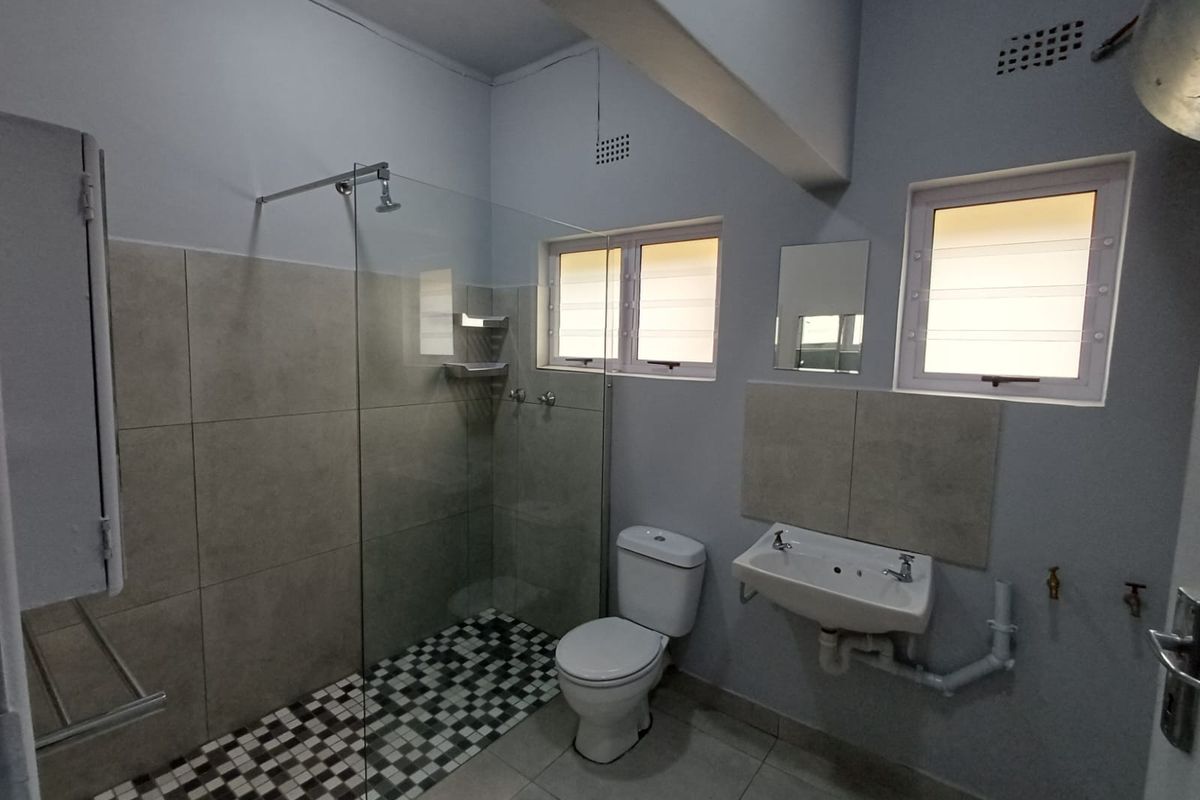 A functional bathroom featuring a walk-in shower with glass screen, wall-mounted basin, and louver windows, finished with neutral floor and wall tiling.