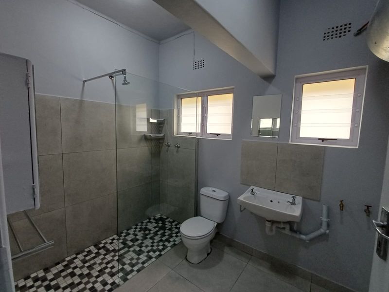A functional bathroom featuring a walk-in shower with glass screen, wall-mounted basin, and louver windows, finished with neutral floor and wall tiling.