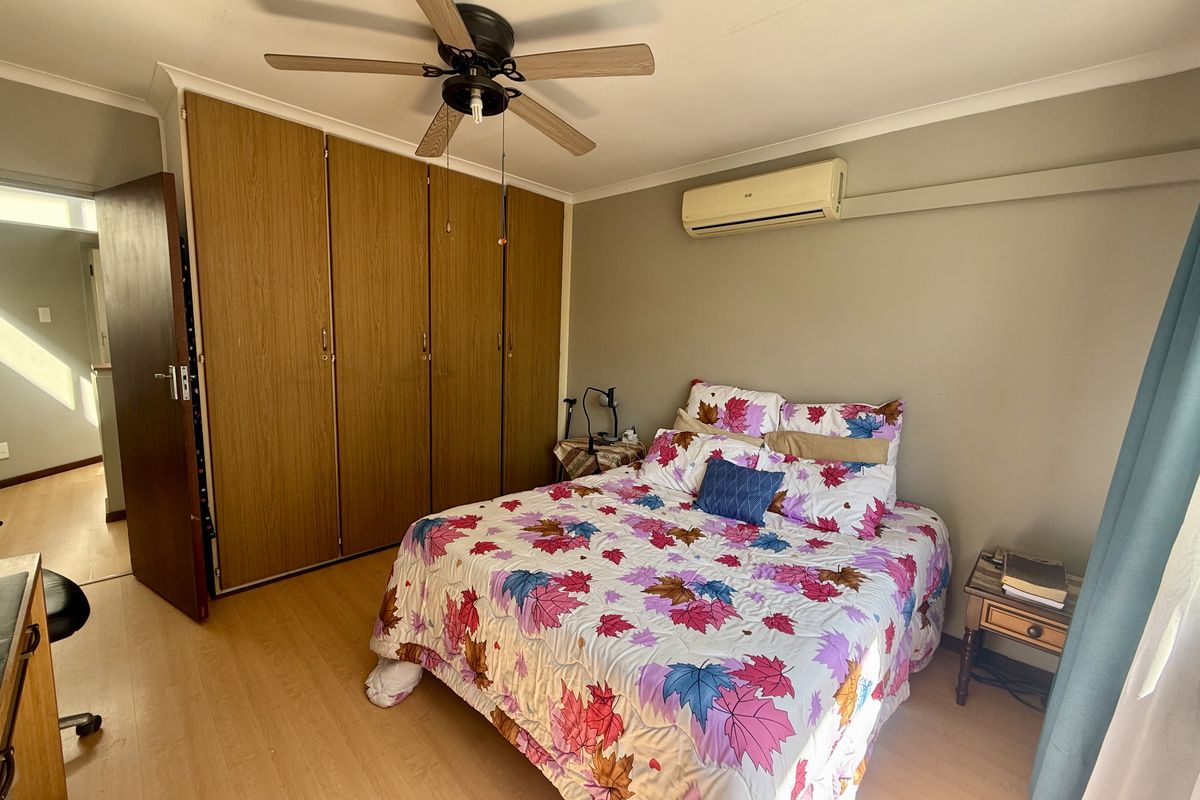 A bedroom featuring laminate flooring, built-in wooden cupboards, a ceiling fan, and a wall-mounted air conditioning unit.