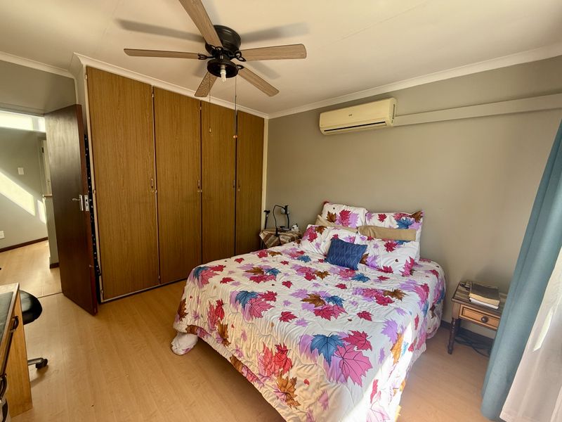A bedroom featuring laminate flooring, built-in wooden cupboards, a ceiling fan, and a wall-mounted air conditioning unit.