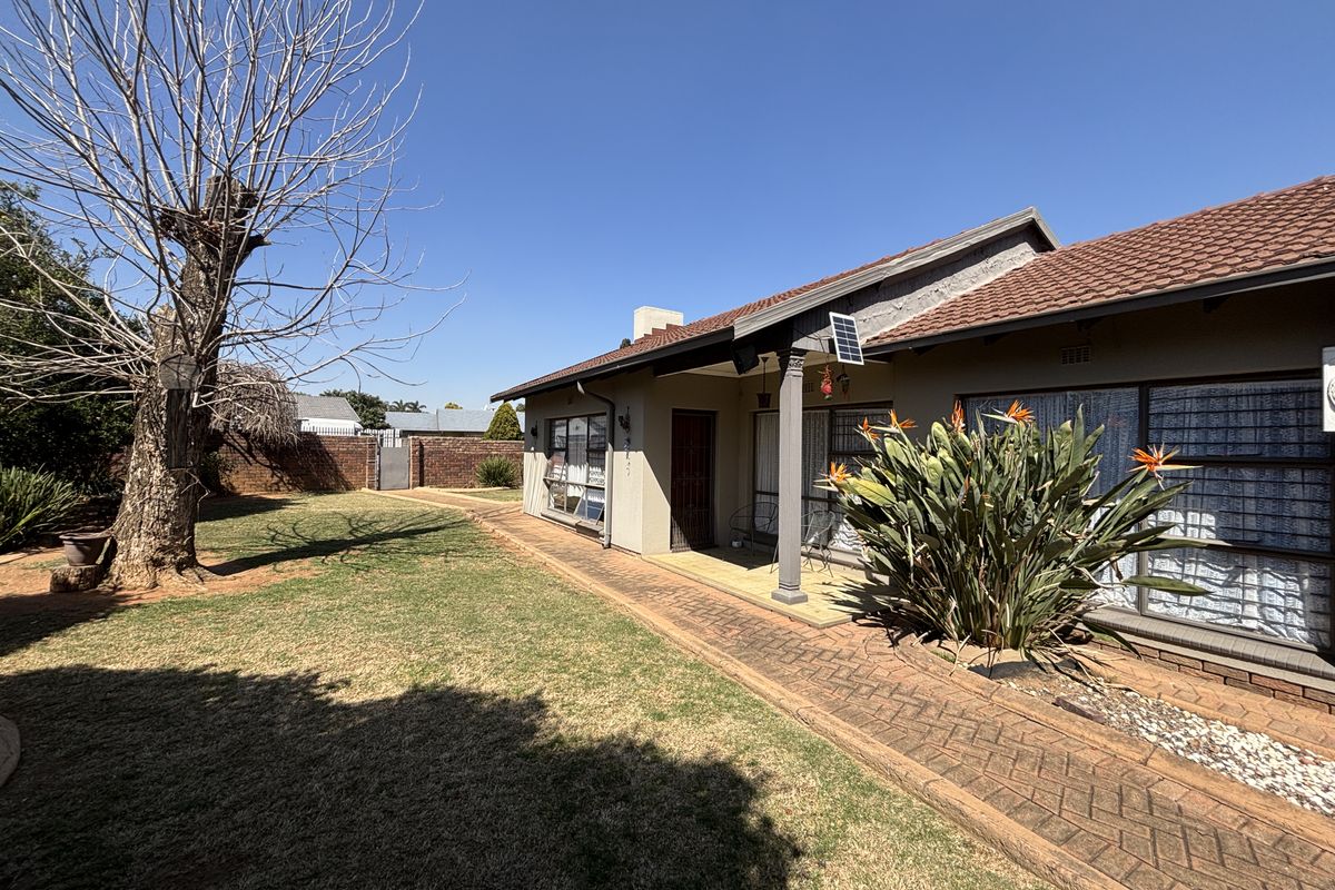 Rear view of the house featuring a covered patio, brick paved walkway, and a spacious garden with established trees and boundary walling.