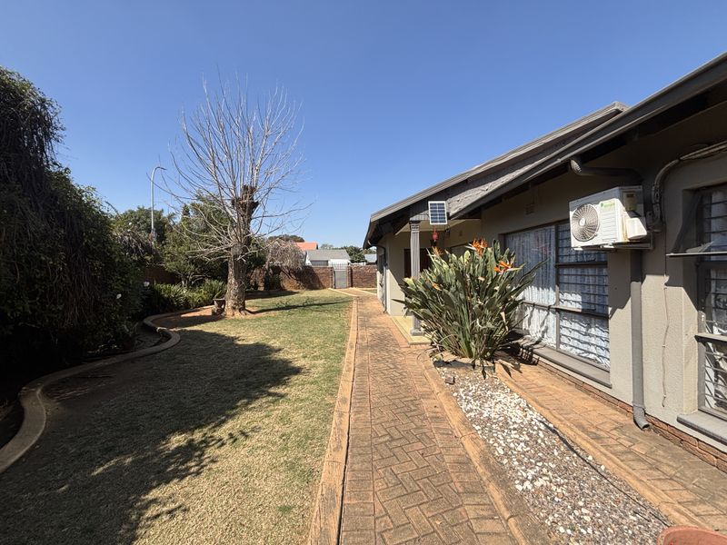 Rear exterior view featuring a brick paved walkway, established garden, and wall-mounted air conditioning unit along the side of the house.