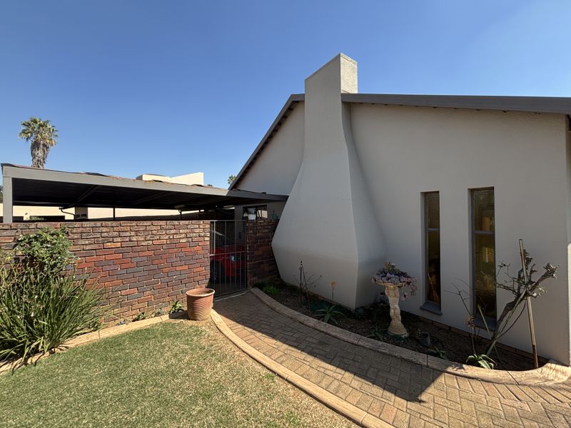 Rear exterior view of the property featuring a paved walkway, brick boundary wall, and a covered carport area.