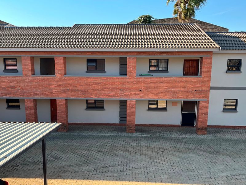 A two-story apartment building featuring a brick and plaster facade, tiled roof, and a paved parking area with a carport.
