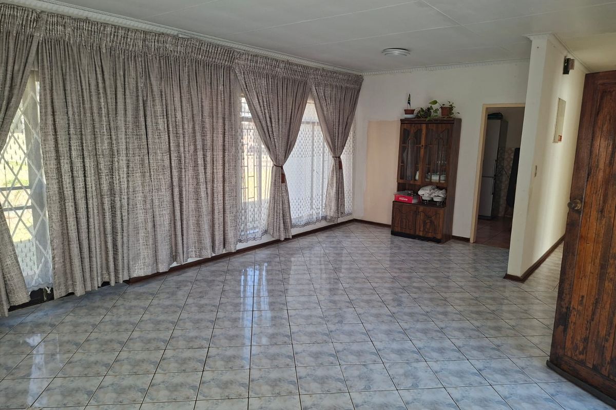 A spacious, light-filled living room featuring neutral tiled flooring, large windows with floor-to-ceiling curtains, and direct access to the exterior.