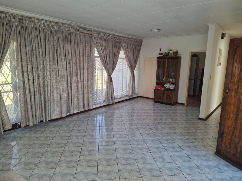 A spacious, light-filled living room featuring neutral tiled flooring, large windows with floor-to-ceiling curtains, and direct access to the exterior.