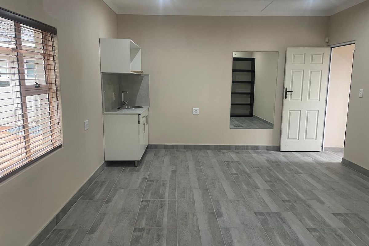 An open-plan living space featuring modern grey tiled flooring, a compact kitchenette with sink, recessed lighting, and a large wall-mounted mirror.