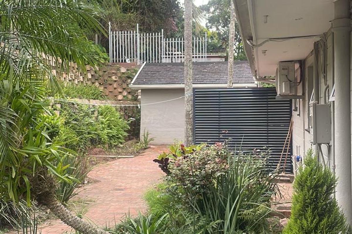 A view of the rear garden area featuring brick paving, established landscaping, a retaining wall, and a secondary structure in the background.
