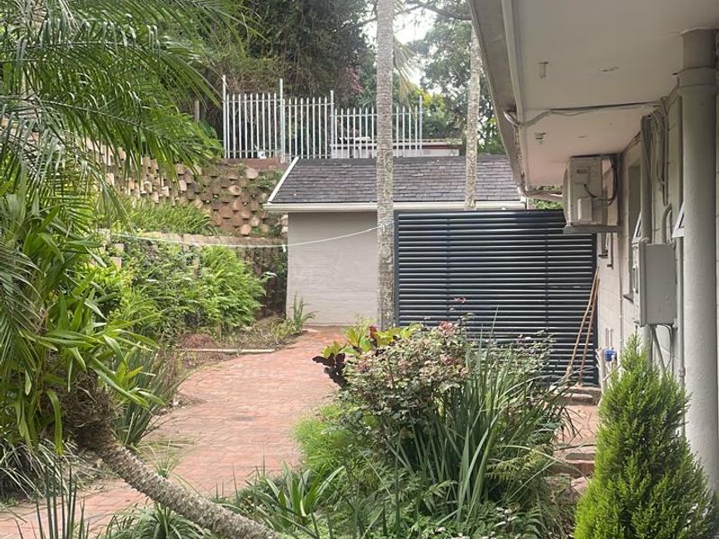 A view of the rear garden area featuring brick paving, established landscaping, a retaining wall, and a secondary structure in the background.