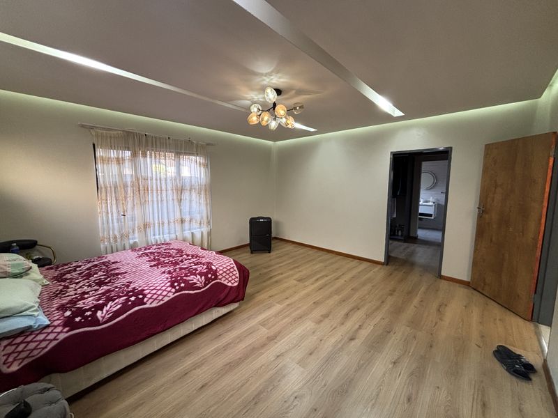 Spacious bedroom featuring light wood laminate flooring, modern recessed cove lighting, and a contemporary chandelier, with an adjoining en-suite bathroom visible.