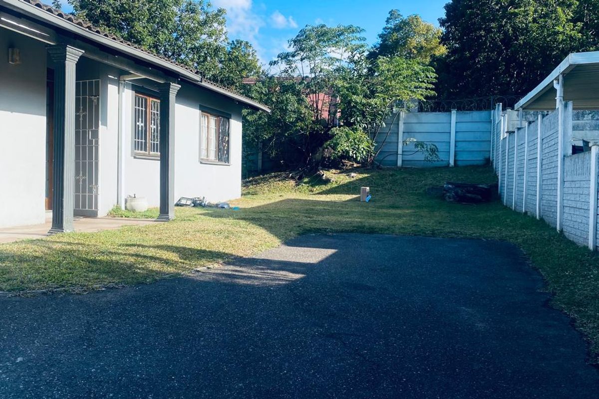 Rear exterior view of the apartment featuring a paved driveway, a small lawn area, and a covered patio with structural columns.