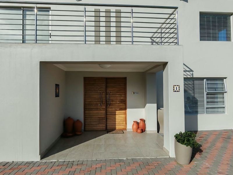 A modern front entrance featuring a double wooden door, a covered tiled porch, and a balcony with stainless steel railings above.