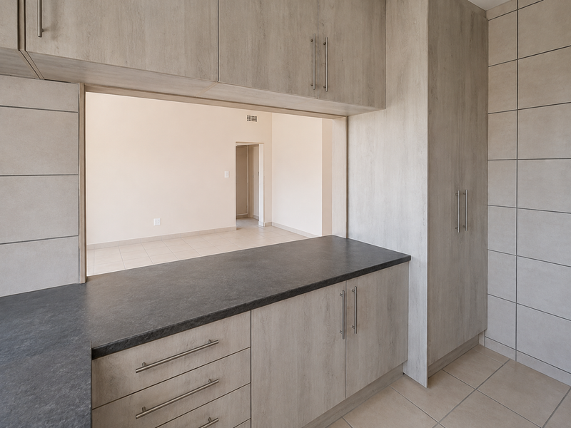 A clean, modern kitchen featuring light wood-grain cabinetry, dark laminate countertops, and neutral tiled flooring, with a convenient pass-through window to the living area.