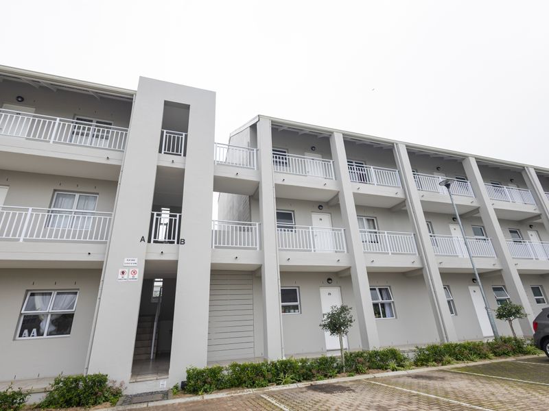 A modern multi-story apartment building exterior featuring clean concrete facades, white balcony railings, and an adjacent paved parking area.