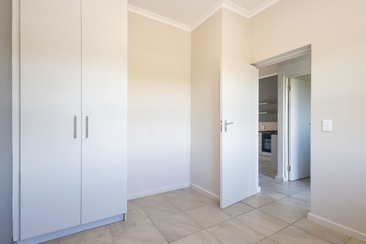 A bright, empty bedroom featuring neutral walls, tiled flooring, and a built-in wardrobe, with a view into the adjacent kitchen area.